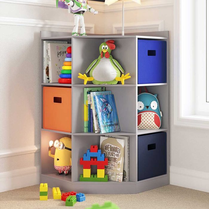 A gray cube shelf displays toys, books, and storage boxes, while a lamp illuminates a playful corner in a bright room.