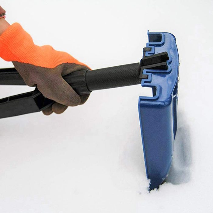 A hand wearing a glove grips a blue snow shovel, which is digging into white snow. The scene suggests cold weather conditions.