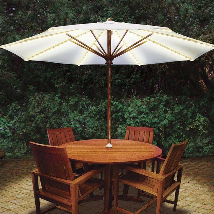 A large, illuminated umbrella stands over a round wooden table surrounded by four wooden chairs, set against a backdrop of green foliage.