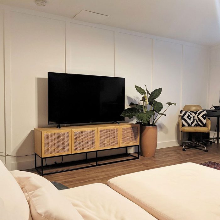 A black television sits on a wooden cabinet, beside a potted plant, in a light-colored room with a brown chair and a patterned pillow.