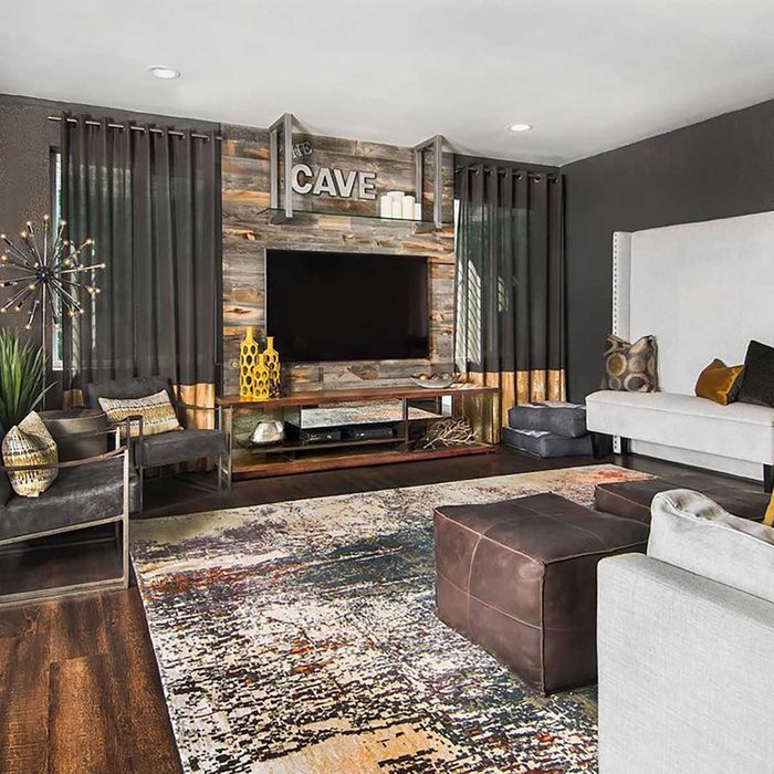 A stylish living room features a central TV unit with a wooden backdrop, surrounded by comfortable seating, decorative pillows, a patterned rug, and dark curtains.