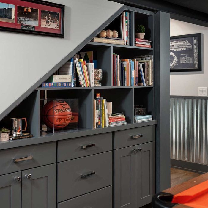 A gray bookshelf holds books and a basketball, displaying a mix of decor and memorabilia, situated in a modern interior beneath a staircase.