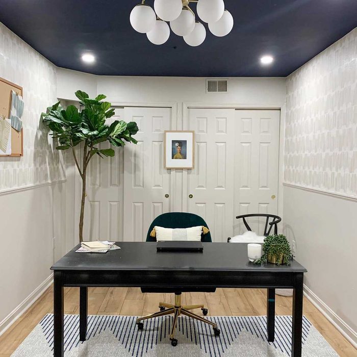 A black desk with a chair sits in a well-decorated office featuring a plant, framed art, and a cozy rug against textured walls.
