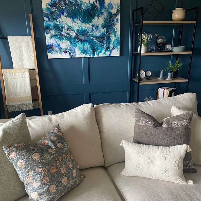 A beige sofa features decorative pillows, while a colorful abstract painting hangs on a dark blue wall beside a bookshelf displaying plants and decor.