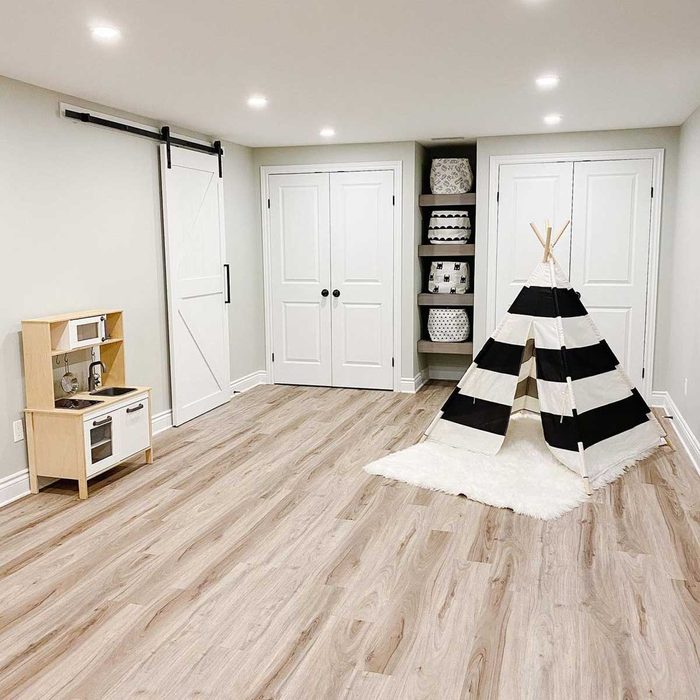 A black-and-white striped teepee stands on light wooden flooring, beside a wooden play kitchen, in a well-lit, neutral-toned room with storage shelves.