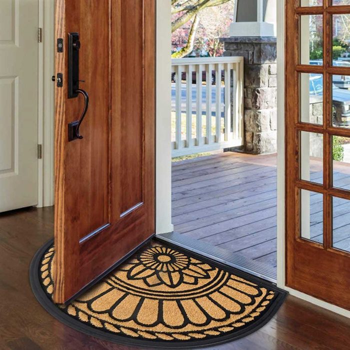 A wooden door swings open, revealing a decorative welcome mat on a wooden floor, with a view of a porch and greenery outside.