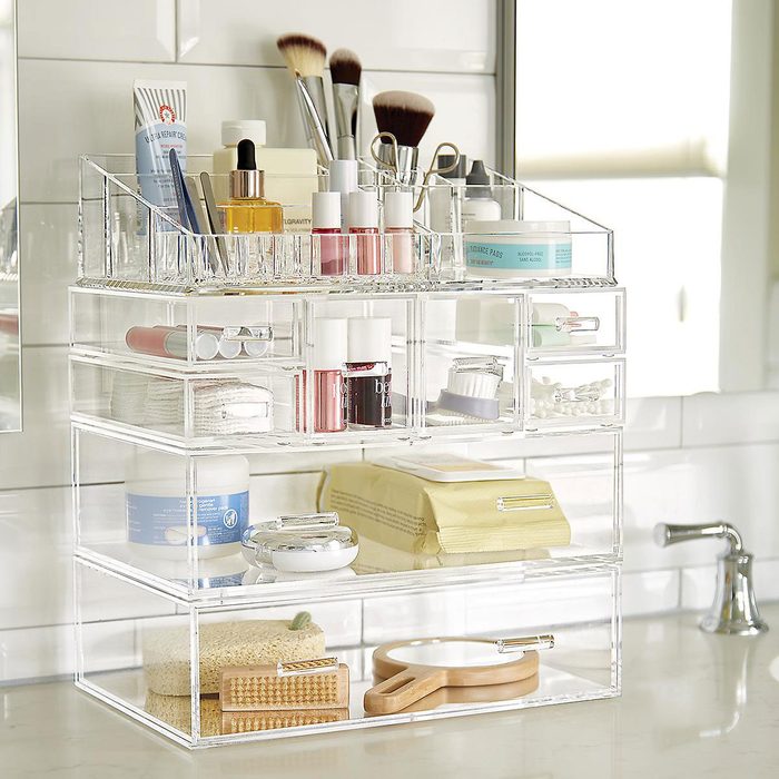 A clear acrylic organizer holds various beauty products and tools, stacked on a bathroom countertop, with sunlight reflecting on the tiles.