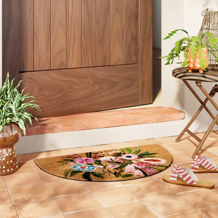A patterned doormat rests on tiled flooring, while a doorway and plants frame a cozy entrance. Nearby, sandals and a small table add to the homey ambiance.