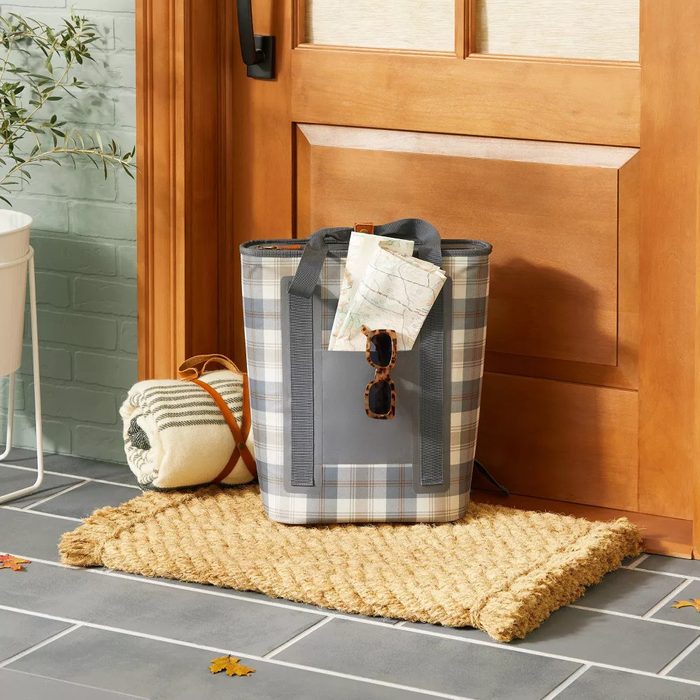 A checkered tote bag sits on a woven mat by a wooden door, holding sunglasses and a map, with a rolled blanket next to it.