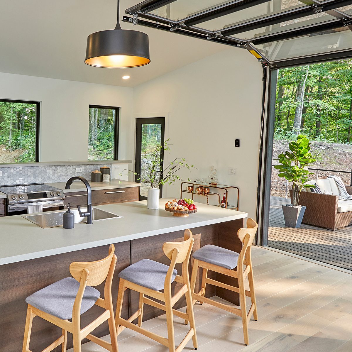 A modern kitchen features a sleek island with wooden stools, a stove, and a scenic view of trees through large windows.