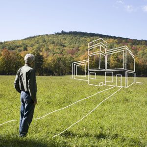 A man stands in a grassy field, observing a white outline of a modern house against a backdrop of trees and a distant hill.