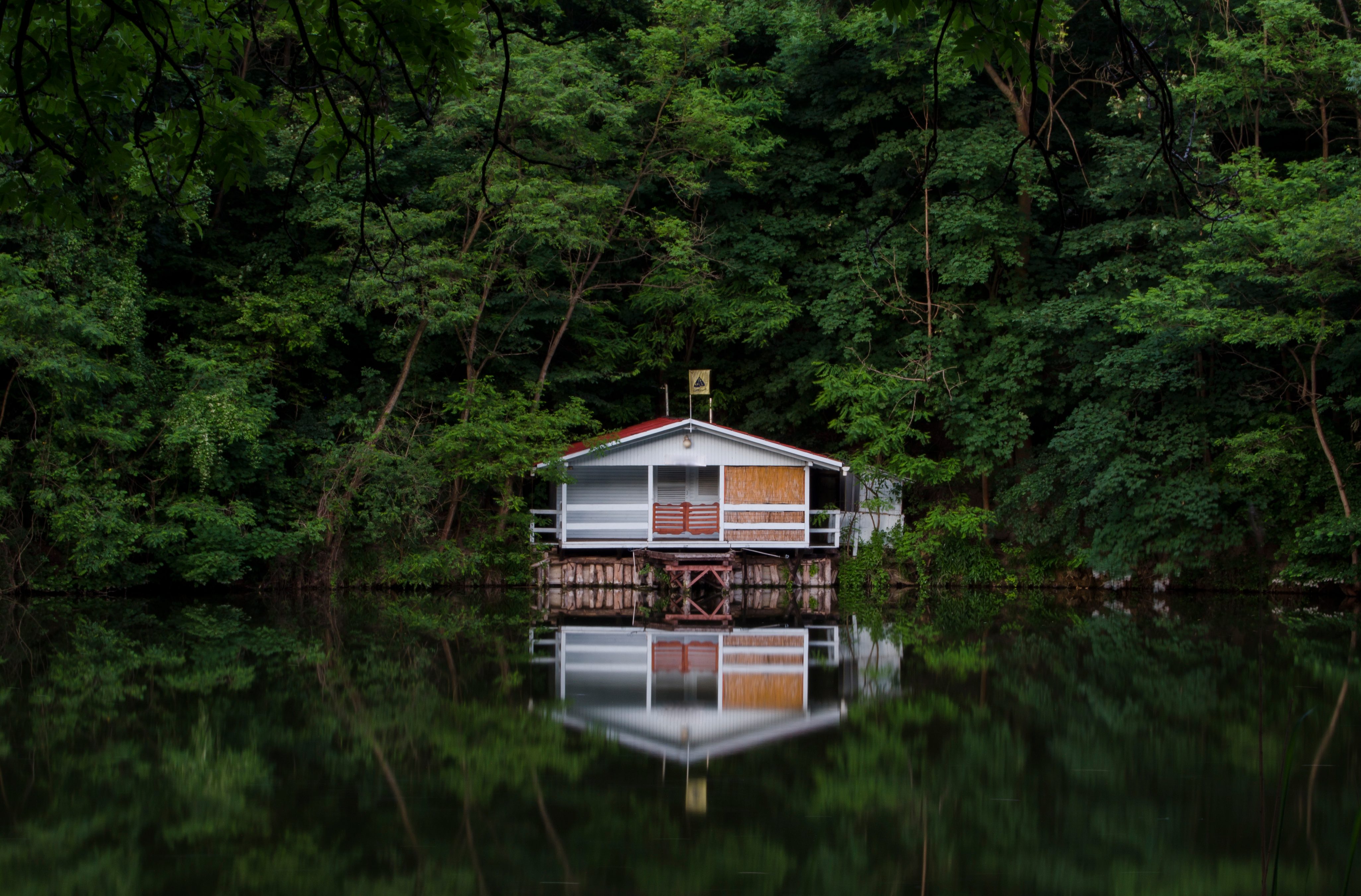 A small, elevated house reflects on a calm lake, surrounded by dense greenery, creating a serene and tranquil atmosphere.