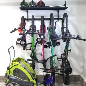 Bicycles hang on a wall-mounted rack, while a green and gray bike trailer rests on the floor below, set against a neutral background.