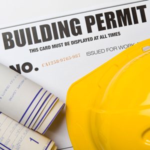 A yellow hard hat rests beside a building permit and rolled blueprint plans, indicating preparation for a construction project.