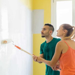 A young man and woman paint a white wall, focused on their task in a bright room with yellow accents and natural light.