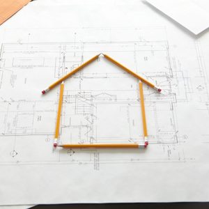 Yellow pencils form the outline of a house on architectural blueprints, which display various floor plans and measurements in a workspace setting.