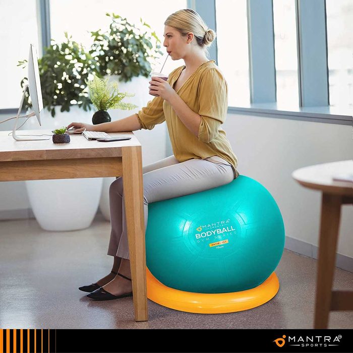A woman sits on a large teal exercise ball at a desk, working on a computer while sipping a drink in a bright, modern office environment.