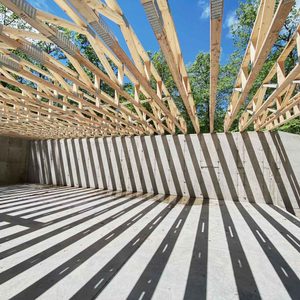 A wooden structure with exposed beams casts long shadows on a concrete floor, illuminated by sunlight filtering through an outdoor environment with trees.