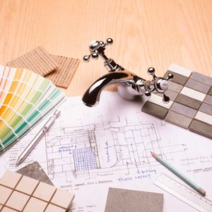 A chrome faucet rests on a wooden surface, surrounded by design plans, color swatches, and tile samples, indicating preparation for a renovation project.