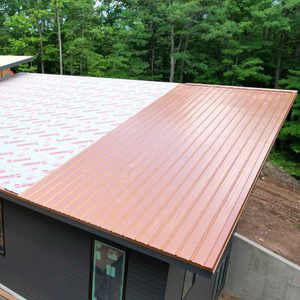 A metal roof covers a structure, with one side completed in copper and the other in lined underlayment, surrounded by lush green trees.