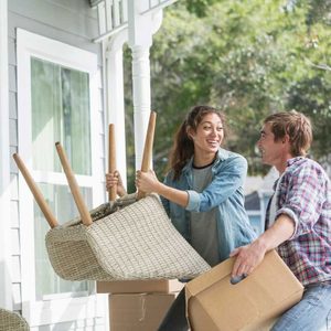 Two people carry furniture and boxes, smiling and engaging in a cheerful conversation outside a house surrounded by greenery.