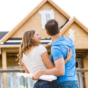 A couple, smiling and pointing, observes a house under construction, holding blueprints as they admire their future home.