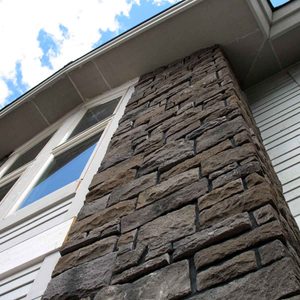 A stone pillar stands tall, showcasing textured, rugged surfaces, with large windows and a blue sky filled with scattered clouds in the background.