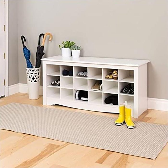 A white shoe storage bench holds various shoes, positioned against a gray wall, with yellow boots on a gray mat and umbrellas in a decorative pot nearby.