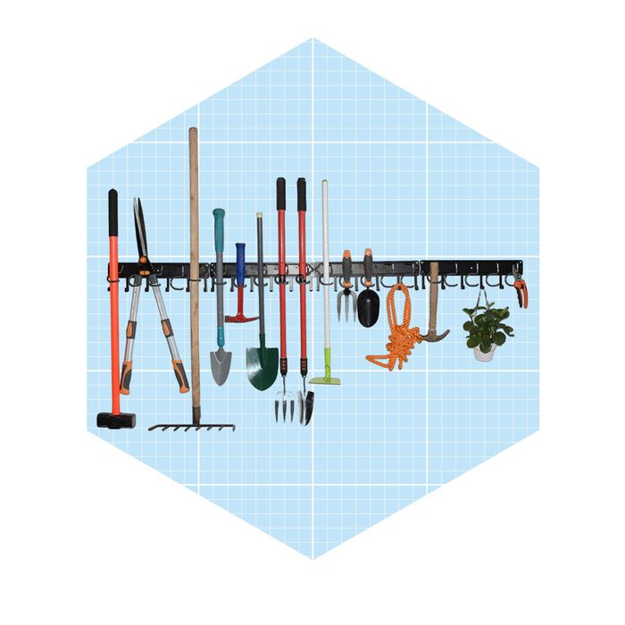 A variety of gardening tools hang on a black wall-mounted rack against a light blue grid background, with a potted plant in the corner.