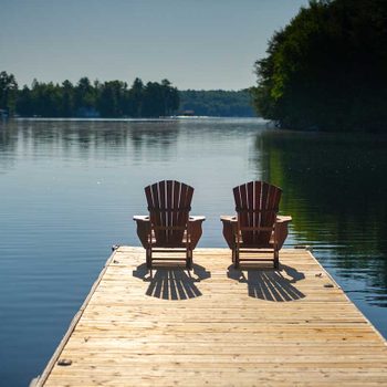 Two wooden chairs are positioned on a dock, facing a calm lake surrounded by trees, casting shadows on the wooden surface under bright sunlight.