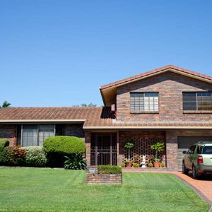 A two-story brick house sits on well-maintained grass, with a driveway leading to a parked vehicle, surrounded by neatly trimmed bushes and colorful plants.