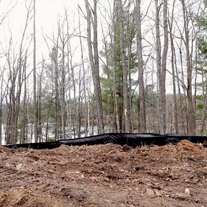 A black erosion control fabric lies on a disturbed, muddy ground, bordered by bare trees and distant green pines, with a body of water visible in the background.