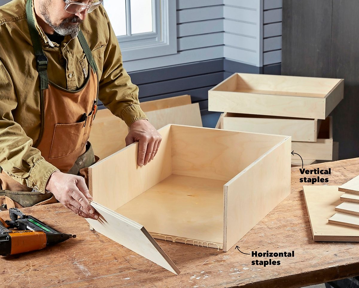 A person assembles a wooden drawer using staples, while several unfinished drawers and tools rest on a workbench in a well-lit workshop.