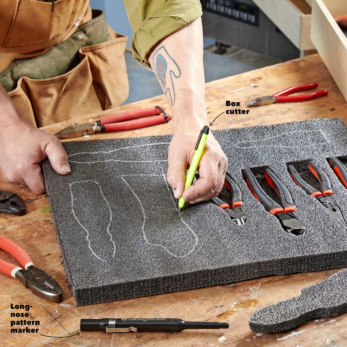 A hand draws outlined shapes on a dark foam pad, surrounded by various tools, including pliers and a box cutter, on a wooden worktable.