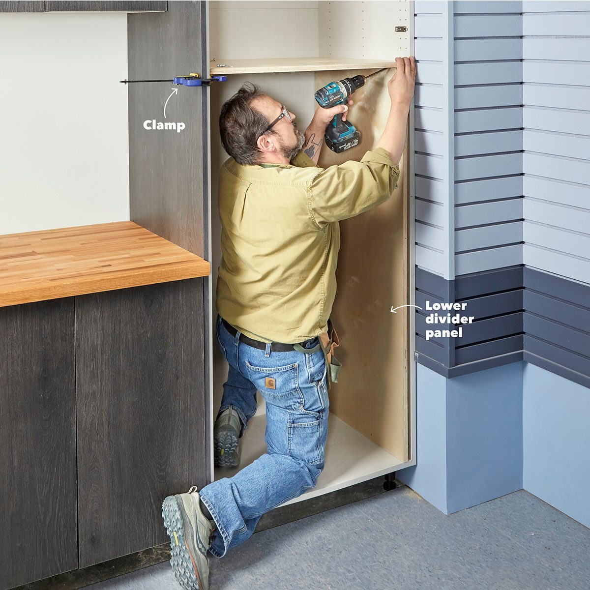 A person drills into a cabinet's interior, securing a lower divider panel with a clamp, surrounded by a kitchen-like setting with a wooden countertop.