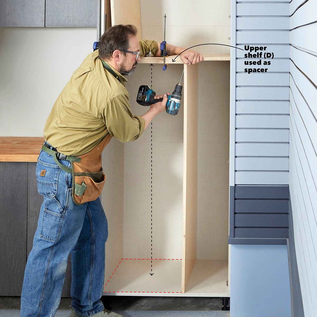 A man uses a power drill to secure an upper shelf inside a cabinet, which is situated against a wall in a workshop setting.