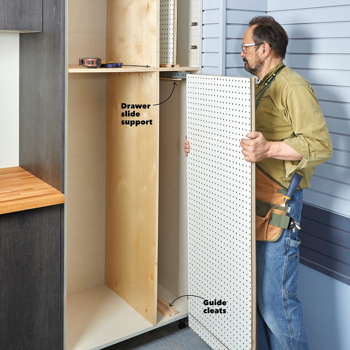 A person is installing a pegboard panel into a cabinet structure, adjusting it near wooden drawer slide supports in a workspace with a wooden countertop.