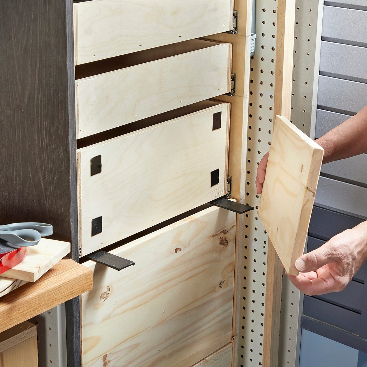 A person is inserting a wooden drawer front onto a cabinet frame in a workshop, with tools and materials visible on a nearby work surface.