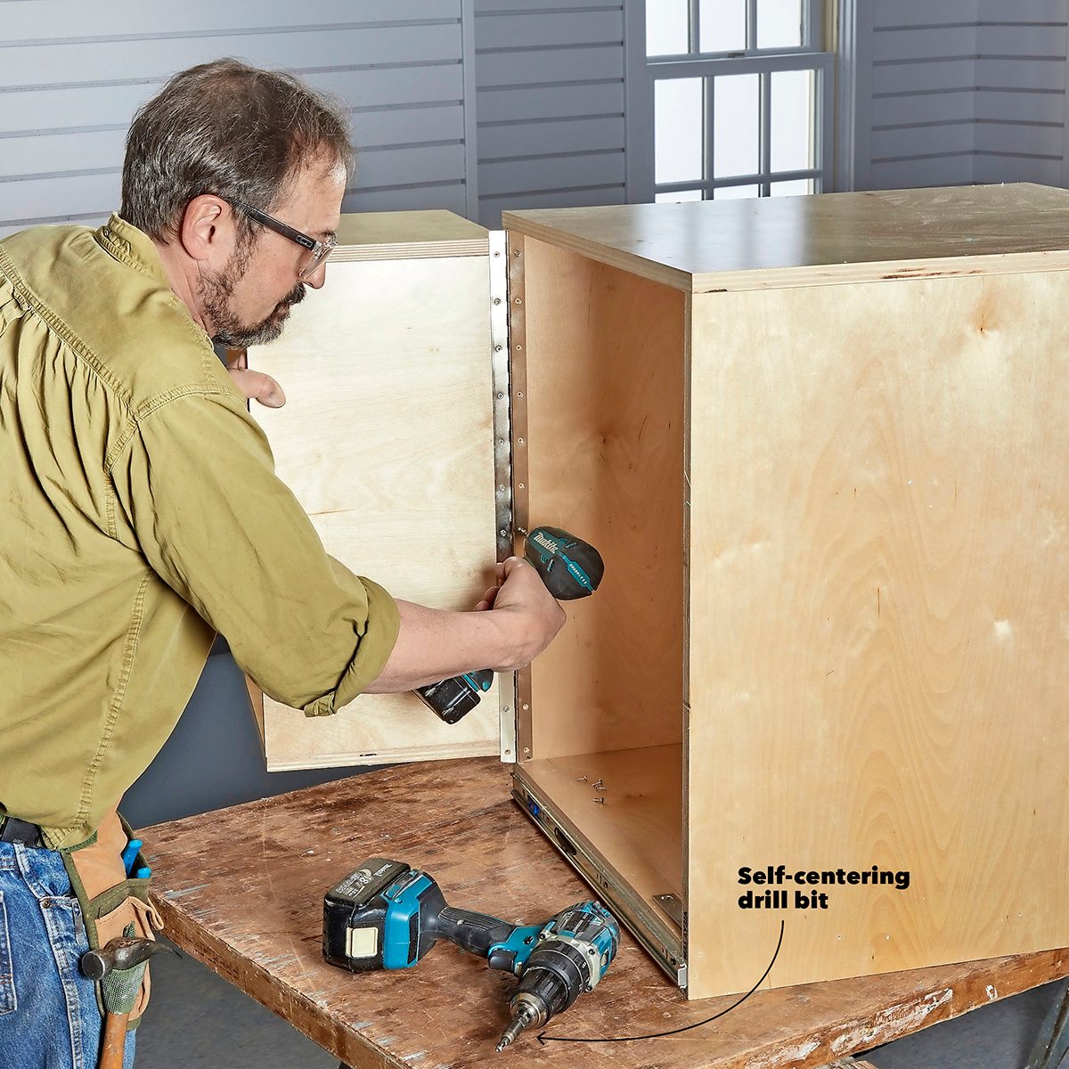 A man drills into wooden panels, assembling them together. Tools lie on a table in a well-lit workshop with a window in the background.