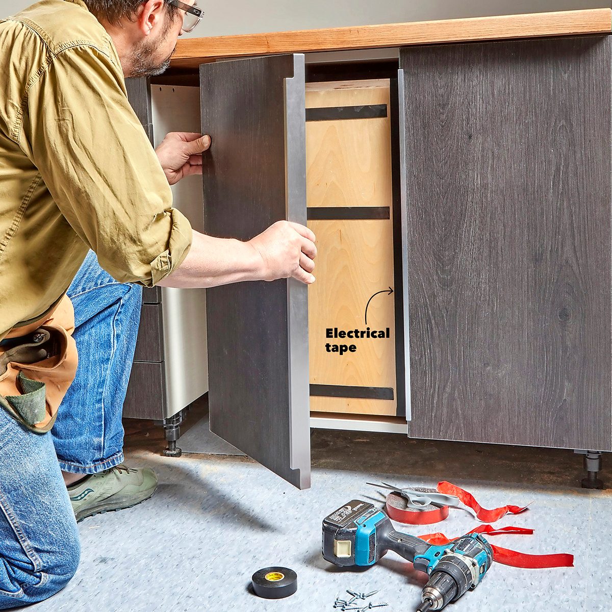 A person is installing a cabinet door, revealing a drawer labeled "Electrical tape" with tools scattered on the floor in a workspace.