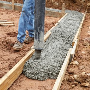 A worker pours wet concrete from a pipe into a wooden form in a dirt area, creating a flat, solid surface for construction.