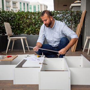 A man assembles white furniture pieces on a wooden deck, using tools and referencing instructions, with greenery and a chair visible in the background.