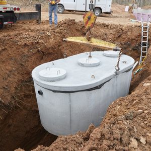 A large concrete tank is being lowered into a dug-out area, surrounded by exposed soil, gravel, and construction equipment in a wooded environment.
