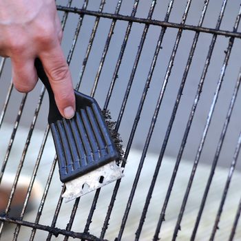 A hand grips a brush, scrubbing a dirty grill grate to remove soot, with a gray concrete surface in the background.