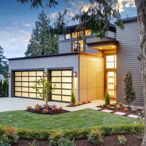 A modern house features large garage doors and a welcoming entryway, surrounded by landscaped greenery and decorative plants under a blue sky.