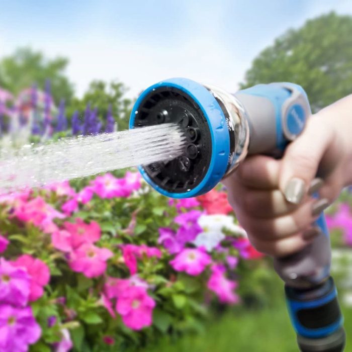 A hand holds a spray nozzle, releasing a stream of water onto colorful flowers in a sunny garden.