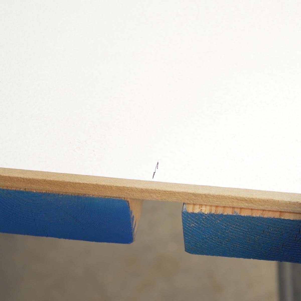 A wooden table edge displays a small crack, while the table is supported by blue padding. The background is blurred, suggesting a workshop setting.