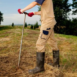 A person in rubber boots uses a digging tool on soil, surrounded by a grassy area and trees, with a cloudy sky in the background.