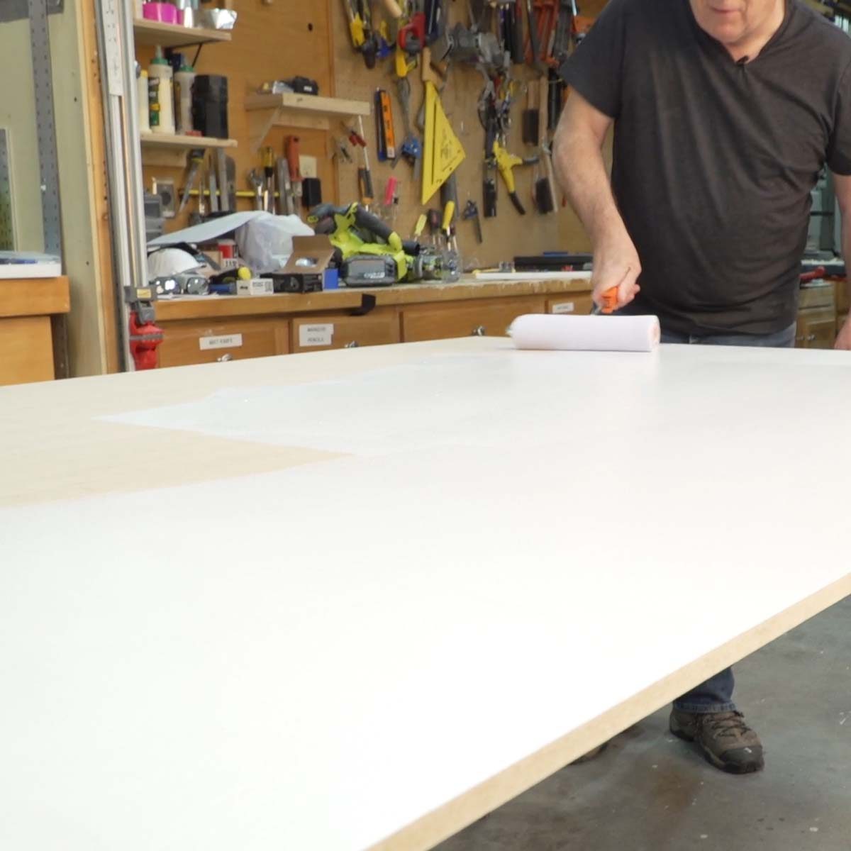 A person rolls white paint on a large wooden surface in a cluttered workshop filled with tools and supplies.