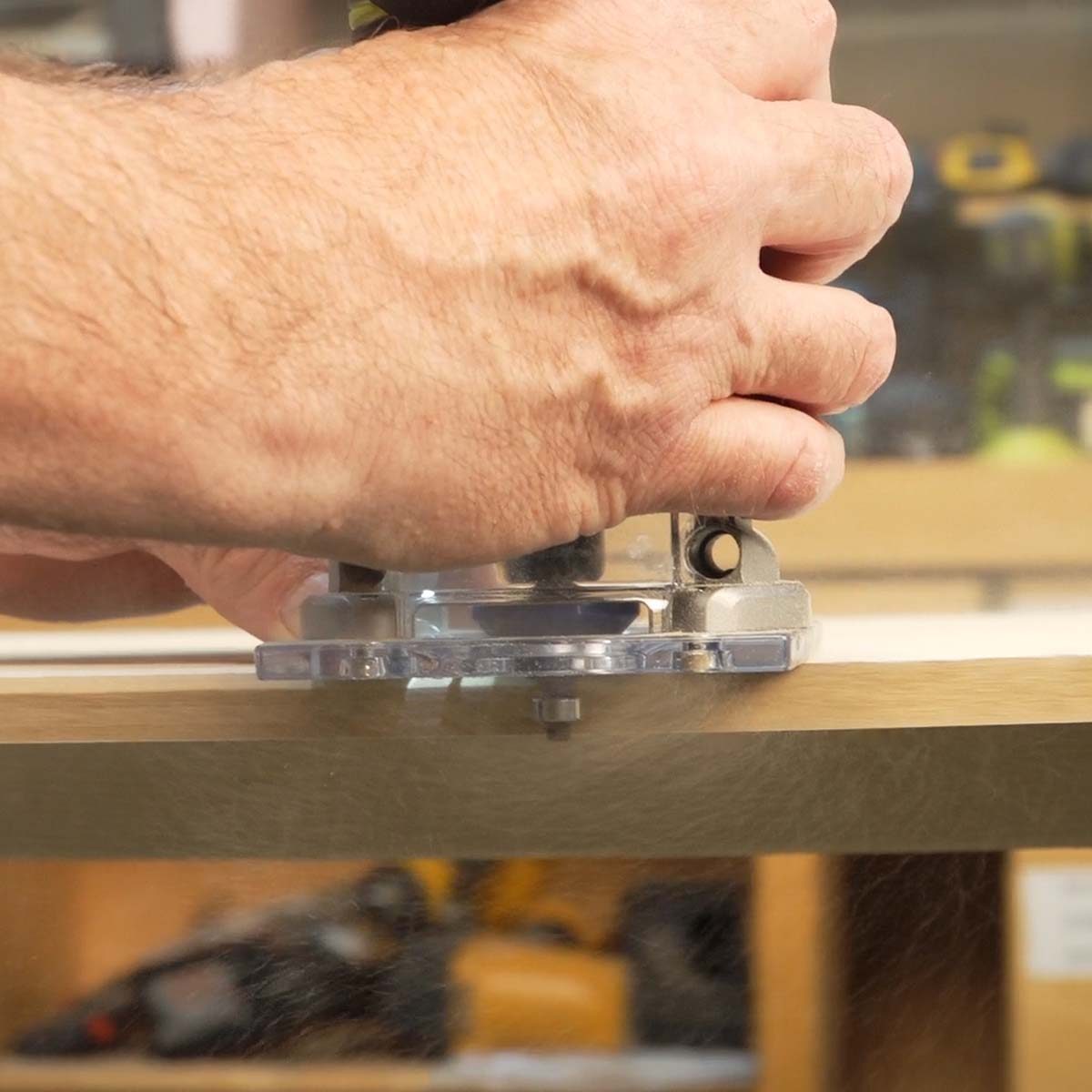 A hand operates a router, shaping wood on a work surface. The setting is a workshop, with tools and equipment visible in the background.